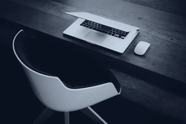 A laptop and mouse on a dark wooden desk beside a white chair