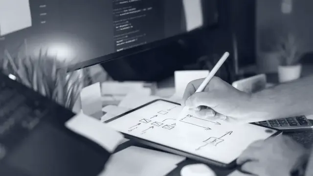 A hand sketching a process flow on a tablet beside a monitor showing code